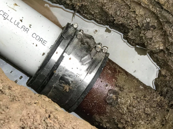 A rubber fernco coupling connects a white PVC pipe to a brown clay pipe in a dirt trench with standing water.
