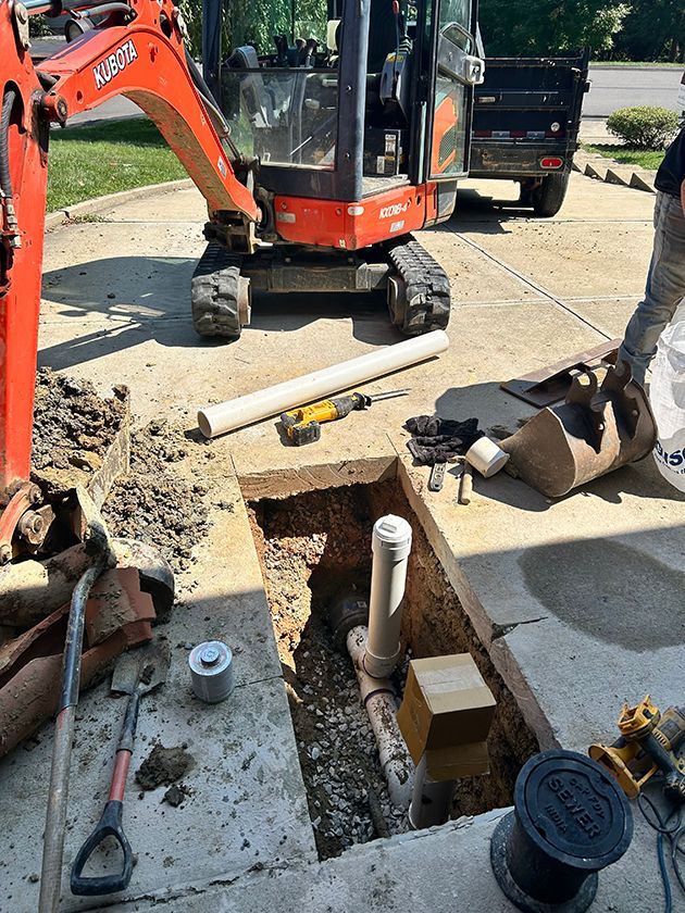An orange excavator is parked near a concrete driveway where a hole has been dug to expose a white PVC pipe.