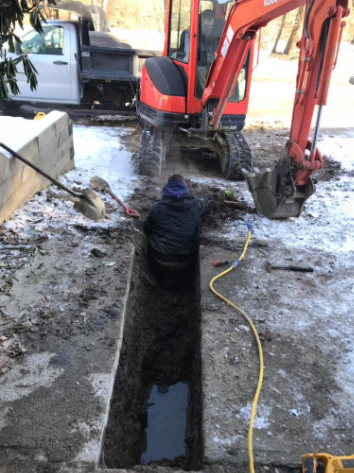 A person kneeling in a narrow trench in snowy ground, with an orange excavator parked nearby.