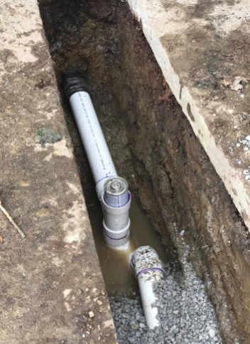 A trench showing gray PVC drainage pipes connected with a vertical cleanout access point surrounded by soil and gravel.