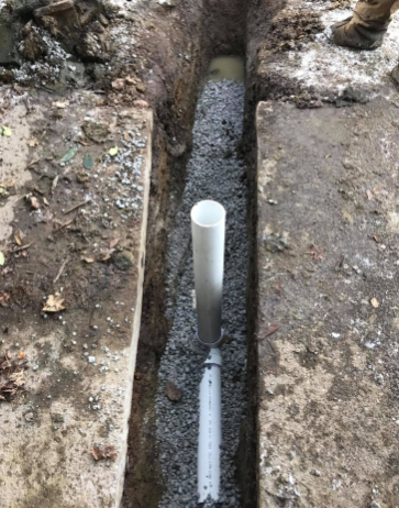 A white PVC pipe assembly with a vertical riser installed in a narrow, gravel-filled trench in the ground.
