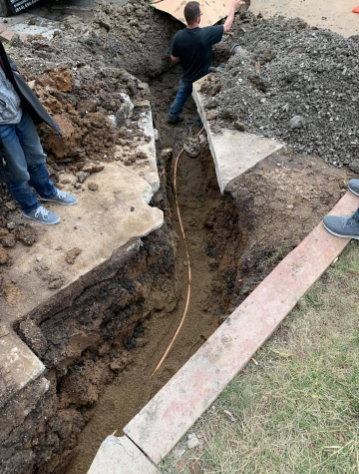 A worker stands in a narrow dirt trench next to an exposed utility line, with observers standing on the concrete edge.