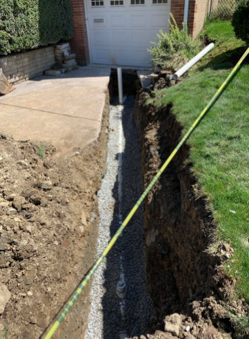 A long, deep trench in a grassy yard containing a drainage pipe surrounded by gravel, leading to a concrete driveway.
