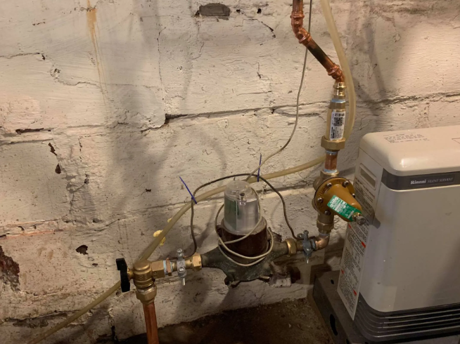 A water filtration system with copper pipes and a control unit mounted against a concrete block basement wall.
