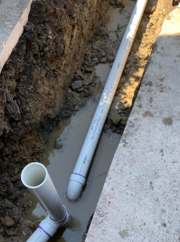 A white PVC pipe assembly with a vertical riser installed in a narrow, muddy trench next to a concrete slab.
