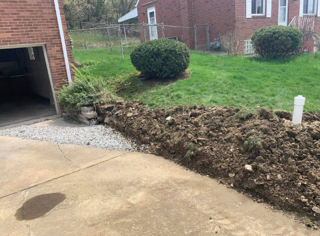 A mound of excavated dirt sits beside a concrete driveway and brick house exterior, with a small shrub and grass behind.