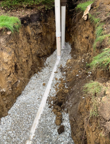 A vertical view of a narrow, gravel-filled trench in the ground containing white PVC pipes connected to a downspout.