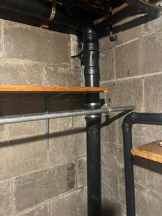 Black plumbing pipes run vertically and horizontally against gray concrete basement walls, with wooden shelves installed.