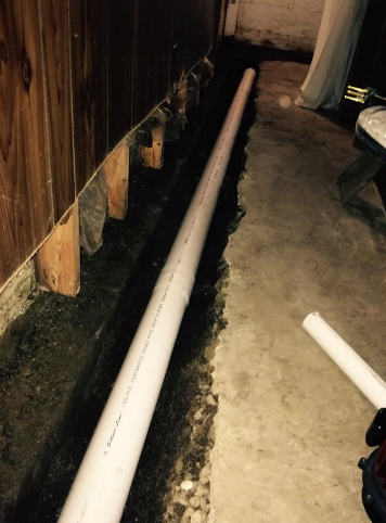 A white drainage pipe installed in a trench dug along the interior wall of a basement concrete floor.