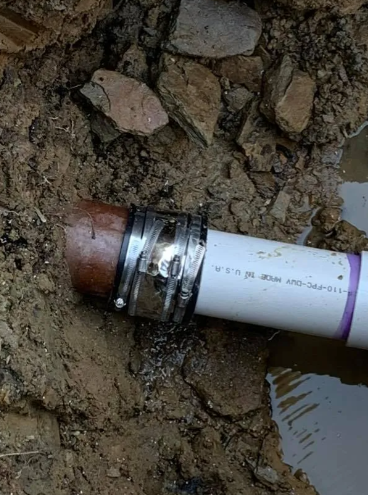 A brown pipe and white PVC pipe joined by a metal shielded rubber coupling in a muddy trench.