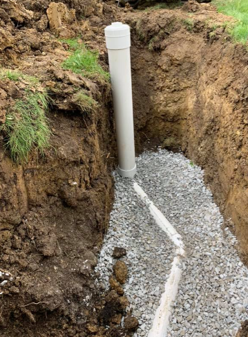 A white PVC pipe stands vertically in a narrow dirt trench, connected to a drainage line bedded in gray gravel.