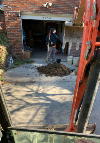 A person with a shovel stands near a pile of dirt in a residential garage doorway, viewed from inside construction gear.