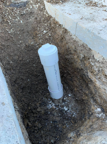 A white PVC pipe capped with a plumbing fitting, extending vertically from the dirt floor of a narrow construction trench.