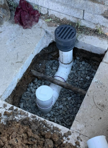 A white PVC drain pipe with a black grate and a capped cleanout sits in a hole filled with gravel next to a concrete slab.