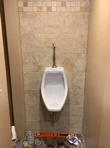 A white porcelain urinal mounted on a tiled wall with a vertical flush pipe and construction tools on the floor below.