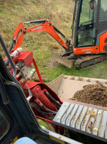 A first-person view from inside an excavator cab, looking toward another orange Kubota excavator working on a grassy hill.