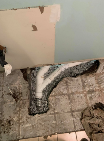 A renovated white PVC drain pipe installed in a concrete floor trench filled with gravel, next to a damaged wall.