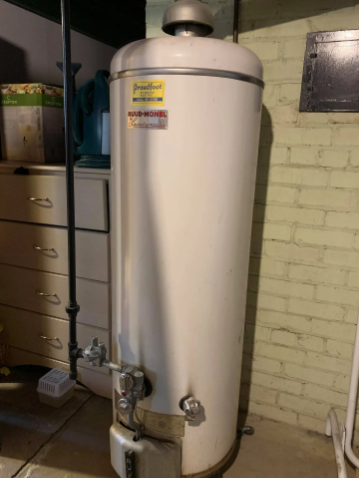 A tall, white, cylindrical gas water heater stands in a basement against a painted brick wall next to a dresser.