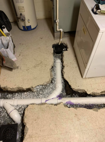 New white PVC plumbing pipes installed in a trench cut into a concrete basement floor near a water heater and appliance.