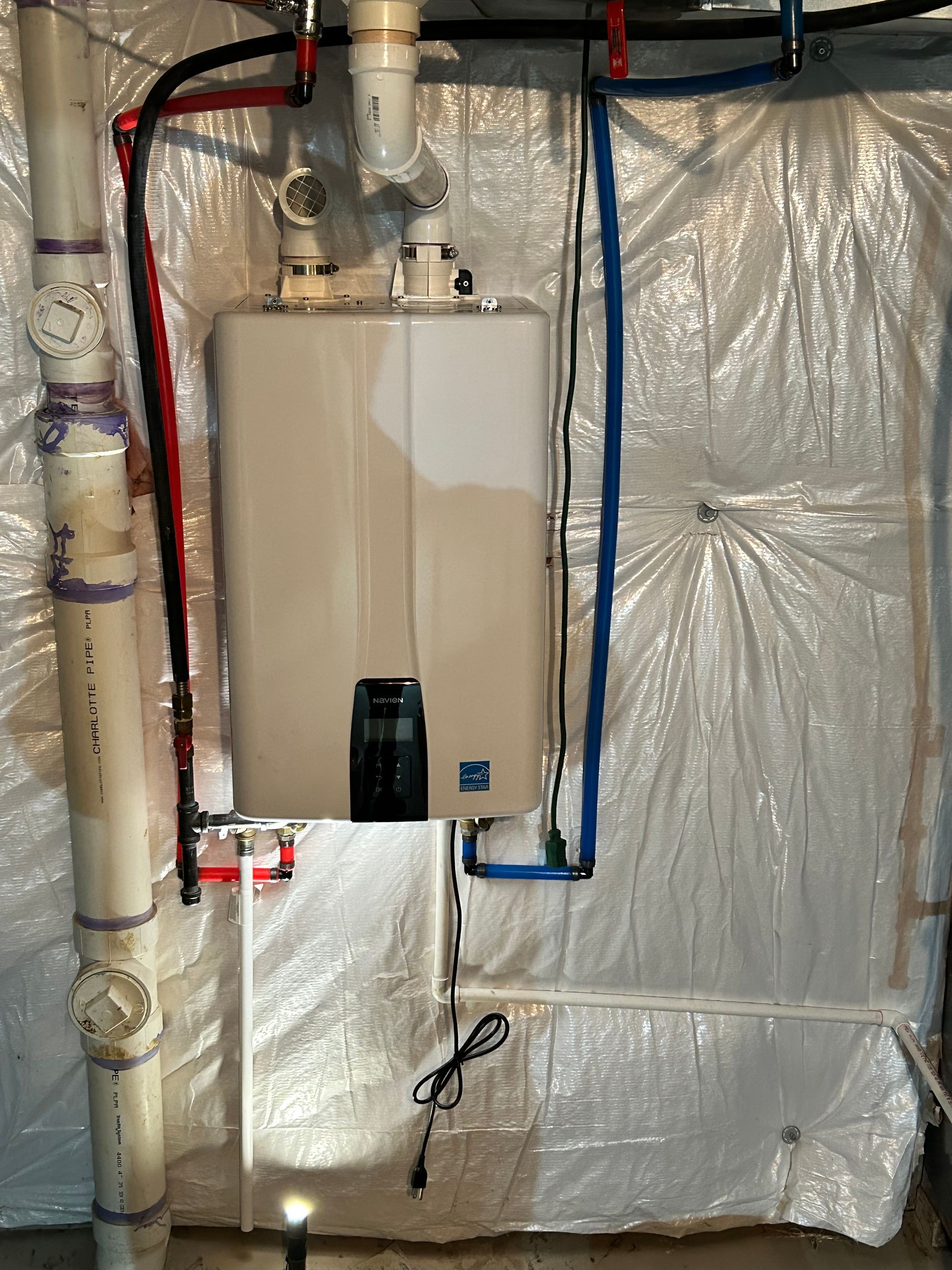 A tankless water heater mounted on a wall with white, blue, and red pipes against reflective, insulated material.