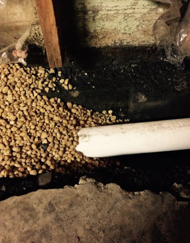 A white PVC pipe discharging water onto a bed of small gravel near a concrete foundation.
