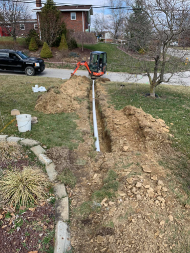 A bright orange mini excavator sits at the end of a long, narrow trench containing a white pipe in a suburban front yard.
