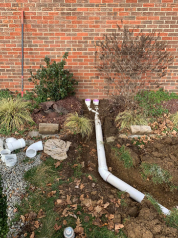 PVC drainage pipes joined into a single line in a trench dug in a yard against a brick wall.