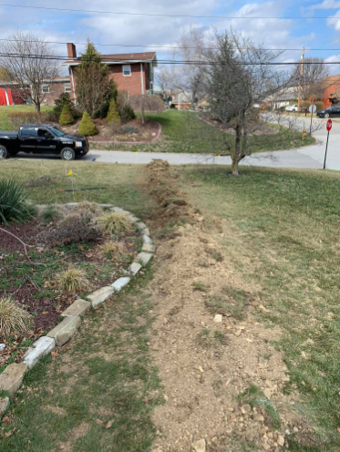 A dirt trench dug through a residential front lawn, situated next to a stone-bordered garden bed and a parked black truck.