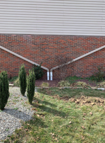 Two gutter downspout extensions run diagonally across a brick house wall, meeting at a central point near the ground.