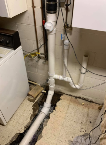 White PVC plumbing pipes installed in a basement, connected to a trench in the concrete floor near a washing machine.