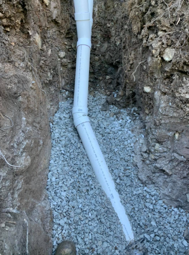 A white PVC pipe assembly laid in a narrow, dirt-walled trench over a bed of crushed gravel.
