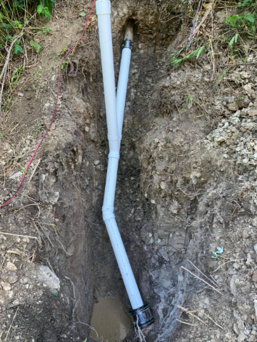 A white PVC pipe assembly with a Y-junction sits in a narrow dirt trench, connected to a darker pipe section at the bottom.