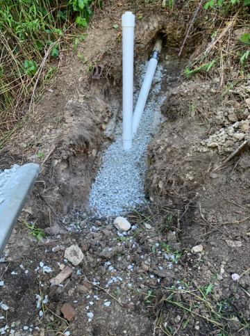 A white vertical PVC pipe connects to a diagonal pipe in a dirt trench partially filled with gray gravel.