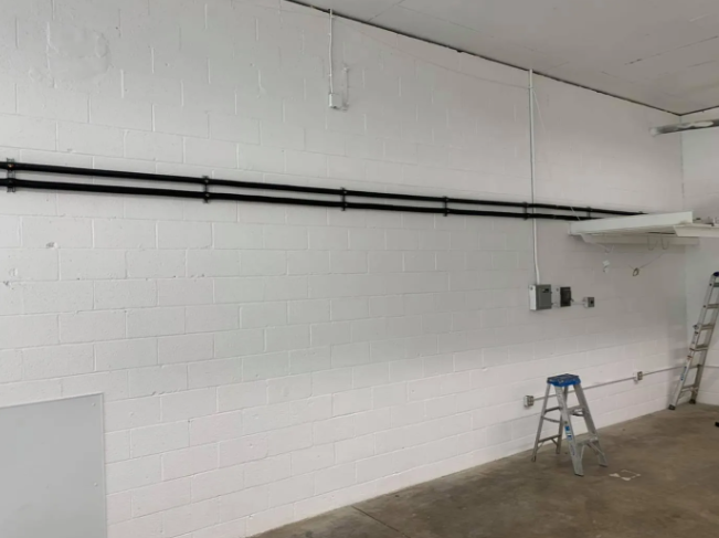 A white cinder block wall with two horizontal black pipes mounted across it, an electrical box, and a folding ladder.