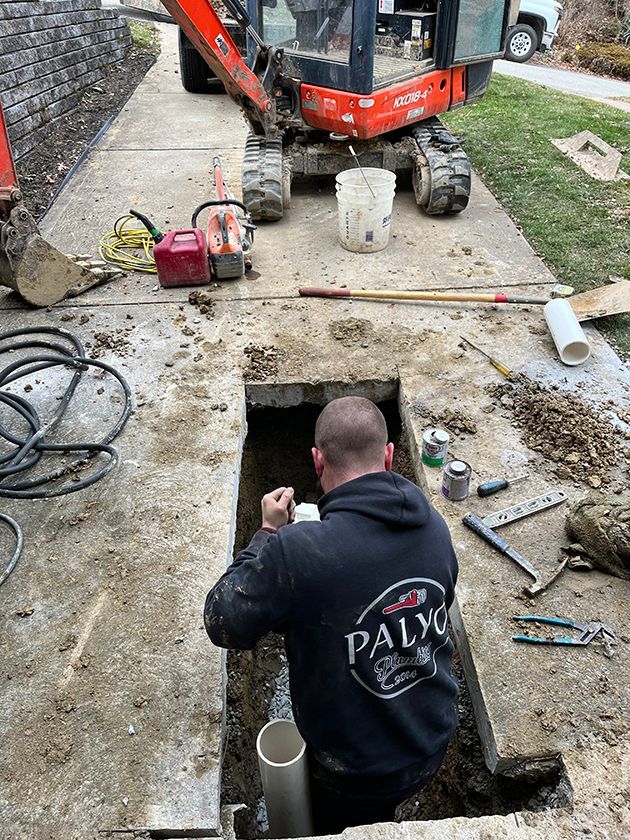 A person in a black hoodie works in a rectangular trench in a concrete driveway near an orange mini-excavator.