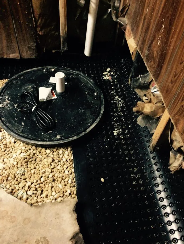 A sump pump basin and drainage matting installed in a basement, featuring gravel, black floor lining, and wall paneling.