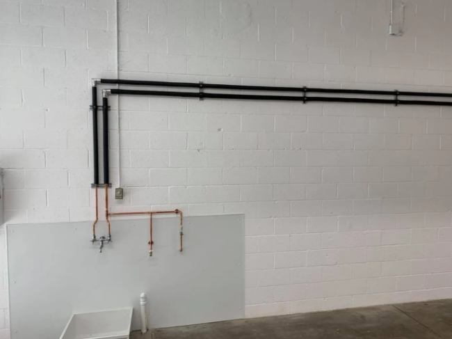 Copper plumbing lines connect to black insulated pipes mounted on a white industrial block wall above a utility sink.