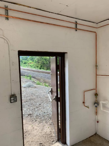 Interior view of a utility room with copper piping along the white walls and an open door showing railroad tracks outside.