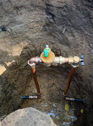 A water pressure regulator and shut-off valve connected to copper pipes, installed in an excavated dirt hole.