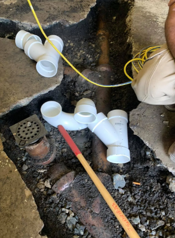 Plumbing repair in an excavated dirt floor, featuring white PVC pipes, fittings, and a drain grate near a worker's legs.