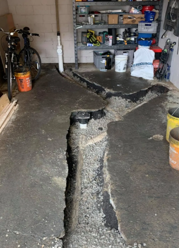 A garage floor with a long, wide trench cut into the concrete, revealing gravel and white plumbing pipes underneath.