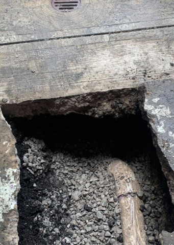 A rectangular hole cut in a concrete surface, revealing gravel and a pipe buried beneath.