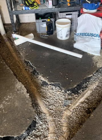 A concrete basement floor with a trench cut into it, showing exposed pipes, with a white PVC pipe assembly nearby.