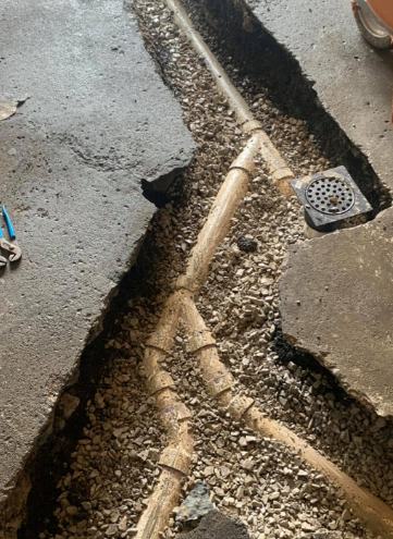 A high-angle view shows an open trench in a concrete floor revealing tan PVC drainage pipes connected to a square drain.
