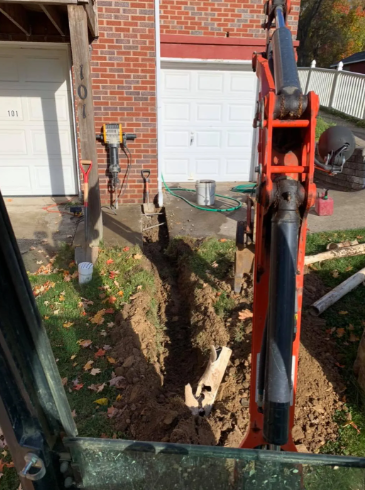 Orange excavator arm reaching into a deep trench dug in front of a brick building with a garage door.