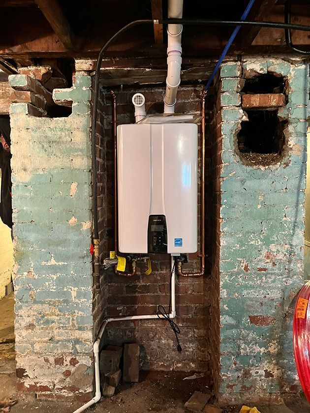 A white wall-mounted tankless water heater installed against a light blue painted brick wall in a basement.