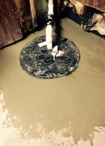 A sump pump basin with a white discharge pipe, set into a floor recently patched with fresh, wet concrete.