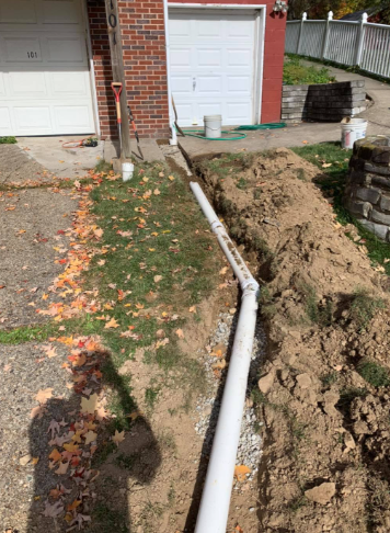 A white PVC pipe runs through a trench alongside a brick house with two garage doors and a pile of excavated dirt.