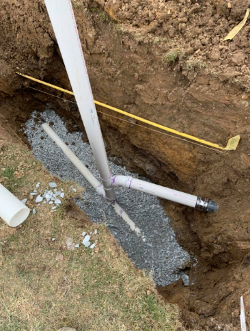 White PVC drain pipes connected at a T-junction, resting on a bed of gravel within a narrow dirt trench.