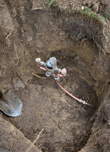 A metal water meter and pipe assembly sit at the bottom of a hole in the dirt, next to a shovel head.